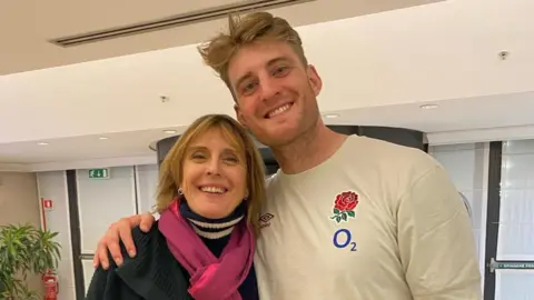 Alex Coles with his arm round his mother, the Reverend Olivia Coles. He is wearing a white T-shirt with the England red rose on the breast. She is wearing a blue jumper and a pink scarf. They are both looking at the camera and smiling.
