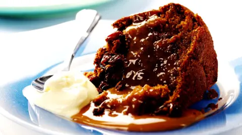 Getty Images A stock image of a sticky toffee pudding on a plate next to a dollop of cream.
