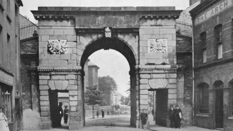 Print Collector/Getty Images/Hulton Archive A black and white archive print of a large stone gate in Derry city centre which forms part of the city's historic 17th century walls.  It has a large, arched entrance and smaller rectangular pedestrian entrances on either side of the arch.  Several people are standing at the gates including a young, barefoot boy who is leaning against the stone. 