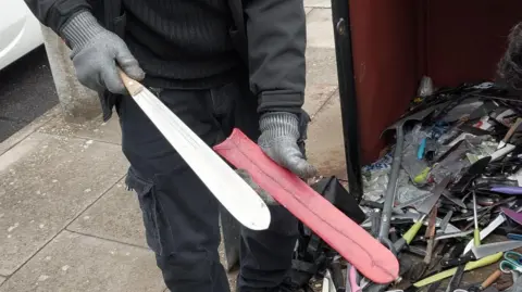 Essex Police A man wearing gloves holds a long blade, which has come out of a red sheath. A large pile of knives are on the floor.