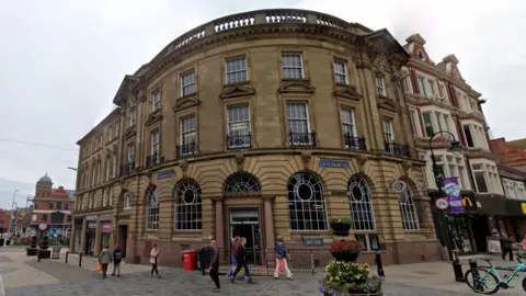 The former Barclays Branch building. The building is on the corner of King Street. It is a curved, sandstone building with many windows. Pedestrians walk on the pavement outside. A McDonald's is next door.