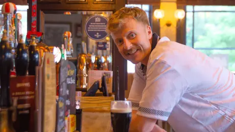 Sam, wearing an England football t-shirt, leaning on a pub bar with a 0% Guinness in front of him while smiling into the camera