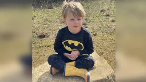 The Menai-Davis family Hugh Menai-Davis sits cross‑legged on a large, flat rock outdoors, wearing a black long‑sleeved top with a yellow Batman logo, matching dark trousers, and yellow Wellington boots. The surrounding area is grassy with sparse vegetation and patches of soil, suggesting a park or rural setting. The child faces slightly to one side with hands resting together in their lap. Natural daylight illuminates the scene, and the background is softly blurred.