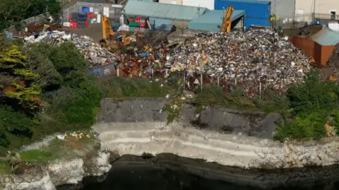 A drone shot showing the proximity between the Guernsey Scrap Metal and Longue Hougue reservoir. Scrap is falling from a large mountain of waste into the quarry. 