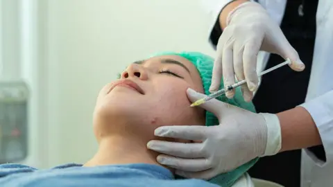 A woman in a blue medical gown and green medical gap lies down with her eyes closed, as a doctor in a white coat and gloves injects a syringe into the woman's face