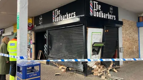 Andrea Pluck/BBC A barber shop with damage to the outside of the store. Bricks and debris are scattered on the floor and an ATM is hanging out of a side of the shop.