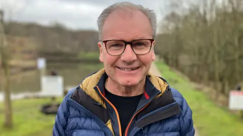 A man with grey hair smiling directly at the camera. He is wearing tortoiseshell glasses and blue puffer coat with a black t-shirt underneath. 