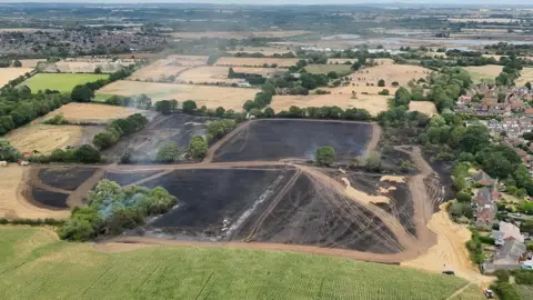 Staffordshire Fire and Rescue Service An aerial image of several fields that are completely blackened and damaged by fire. There is white smoke rising from them. On the right, the charred field reaches right up to the boundary of some houses