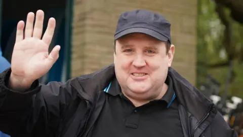 PA Media Peter Kay stands in a street wearing a black jacket and cap. He is waving at the camera. 