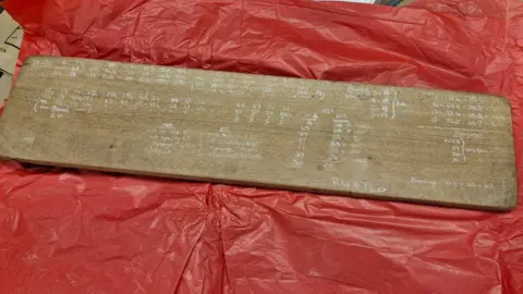 Wolverhampton City Archives A wooden board used for marking fermentation data. It has white unreadable writing on it and it sitting on red tissue paper.