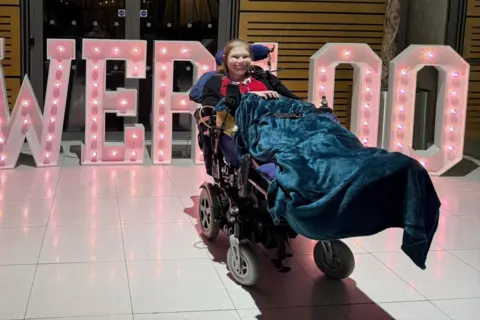 Lucinda Ritchie Woman with dark blond hair lies slightly reclined in a long wheelchair with her legs out long, covered in a dark blue blanket. She is smiling. She is wearing a red top and has an oxygen tube. Behind her are huge pink lit up letters, spelling POWER 100.