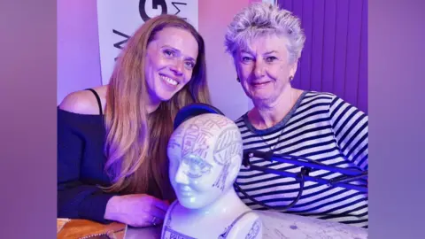Sheila Hannon Rose Wallis, left, with long ginger hair, smiling. Sheila Hannon is right, an older lady with cropped grey hair and a black and white striped top. They are sitting in front of a microphone pop filter, and a phrenology skull