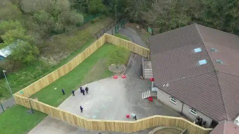 The image shows an aerial view of a school site bordered by tall wooden fencing that forms a zig‑zag shape around a section of playground and green space. The building sits to the right of the frame with a brown tiled roof and several skylights. The fenced‑off area creates a private outdoor space where a handful of pupils and a few adults can be seen standing or walking across the playground. 