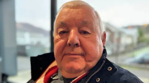 A man sitting down with short grey hair wearing a navy coat and scarf 