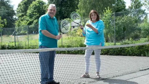 Rugby Borough Council A man and a woman pose, holding tennis rackets behind a net while stood on a grey tennis court