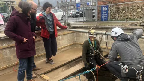 Rejoicing in Ripon Blog/Tim Flanagan A group of five people gathered around a large hole in the ground, with three standing and observing while two helmeted workers in harnesses prepare ropes and equipment for a descent.