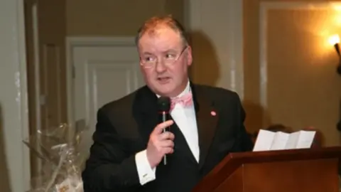 Balding man with clear glasses holding a black microphone and is in a black suit with a white shirt underneath and a pink bow tie.