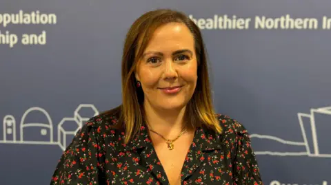 A close-up image of Rachel Spiers standing in front of a dark background with some text written in white. She is a young woman with straight, brown hair at shoulder length and she is wearing a floral patterned dress and a gold necklace. She is smiling at the camera with her mouth closed.