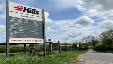 Environment Agency Signage for the landfill site against a brown wooden fence on the side of a grassy verge with wild flowers. It is adjacent to a country road.