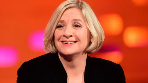 Ian West/PA Wire Comedian Victoria Wood appearing on the Graham Norton Show in 2014. She has a blonde bob haircut and is wearing a black blazer. The background is bright red, with purple and orange lights.