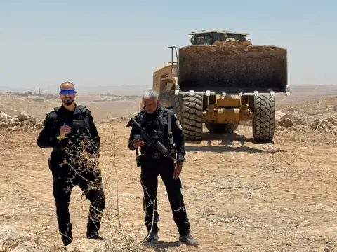 Oren Rosenfeld/BBC Police guard a digger extending the settlement of Carmel near Umm al-Khair, in the southern West Bank