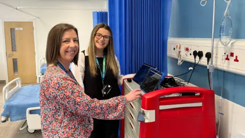 Louise Wood and Georgia Stevens are using two tablets on a teaching hospital ward. They are both smiling at the camera. There is a hospital bed behind them.