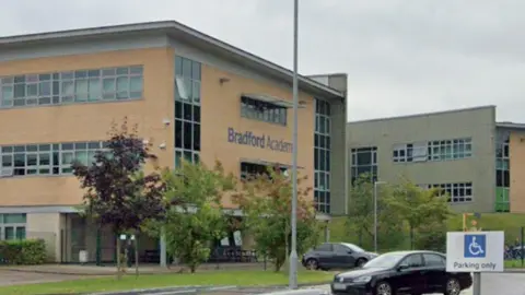 Google A large brick-built school with green and red trees outside and some cars parked in the car park