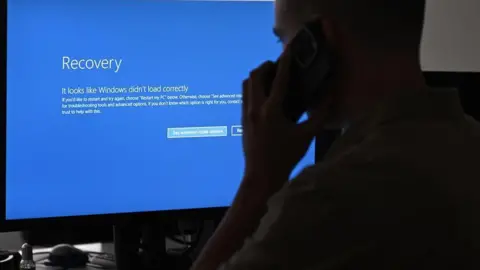 Getty Images A man looks at a computer screen, which shows a blue screen with a sad face emoji. It is an error screen showing a computer has crashed