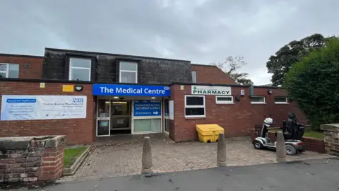 The exterior of one of Drs Reddy and Nunn's GP surgeries in Bridlington. It is a two-storey red-brick building with a blue sign reading "The Medical Centre", and a white sign reading "Pharmacy". A mobility scooter is parked outside.