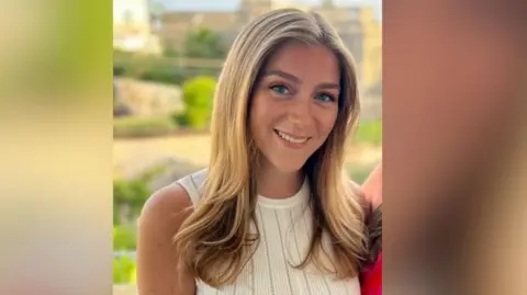 Hannah Dyos smiles at the camera wearing a white dress, she has long blonde hair.