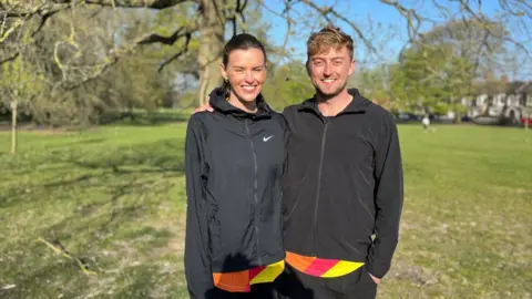 A woman and a man smile while standing together in a park - both are wearing black sports jackets