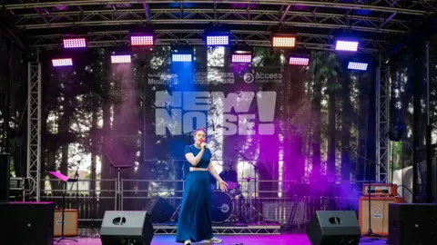 A woman standing in the middle of a stage. She is wearing a blue top with matching blue trousers. She has ginger hair and is holding a microphone to her mouth. Behind her, the stage is lit with purple and blue lights. There is a drum kit, and several stands and speakers. 