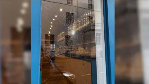 A window pane has been smashed. Behind the window is a shop floor with low level cupboards running down the right and spot lights shining down from the ceiling.