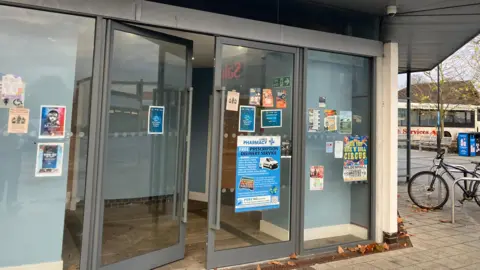 Vikki Irwin/BBC A close-up of floor-to-roof metal-framed glass doors, with a number of posters and flyers stuck to them. Above is a jutting-out roof ledge. To the right, a bike tied to a bike rack is just in shot, and beyond it there is a white single decker bus
in the distance.