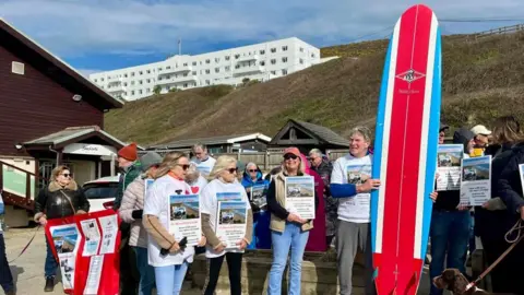 People gather on a beach holding protest posters. One man is holding a red, white and blue surfboard. 