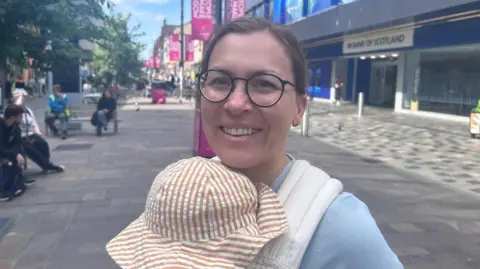 A woman carrying a baby. She is wearing a blue jumper and glasses and the baby has on a striped hat.