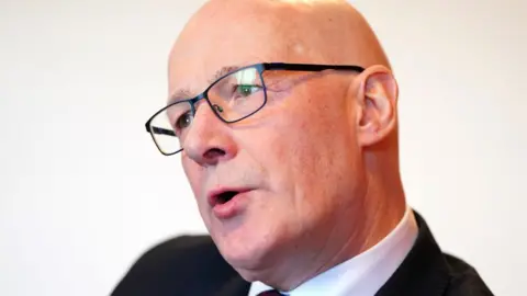 PA Media John Swinney, who is bald and wearing glasses, in a close-up shot against a white background. 
