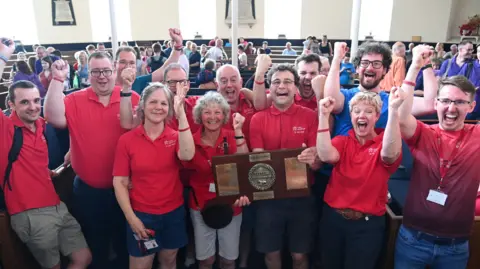 Roger J Lawson A group of people wearing matching red shirts stand closely together inside a large hall with wooden seating. Several individuals raise their arms, and one person at the centre holds a large wooden plaque with metal plates. In the background, more people are seated and standing among rows of benches, with tall windows and bright indoor lighting.