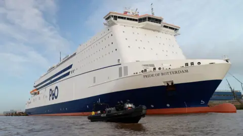 A large white and blue P&O ferry, named Pride of Rotterdam, towers above the water as it moves or sits alongside the river. In the foreground, a small black police rigid‑hulled inflatable boat travels across the brown, choppy surface, appearing tiny in comparison to the huge ferry. Several officers in dark protective gear are on board the smaller vessel.