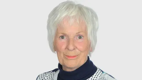 A woman in her seventies wearing a roll-neck blue-and-white top smiles at the camera.