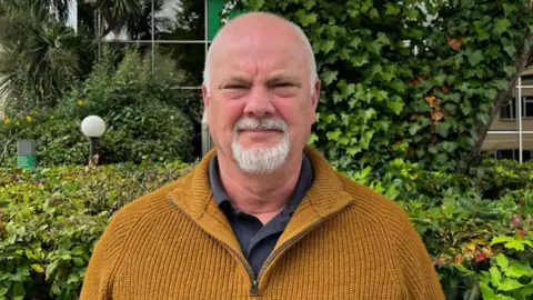 BBC Andy Gealer is wearing a mustard coloured fleece stands in front of some bushes and is looking into the camera