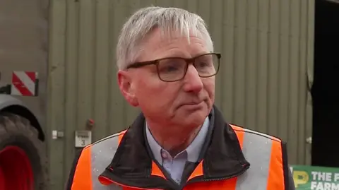 A man looks to the right of the camera. He has short grey hair and has glasses. He is wearing an orange hi-vis jumper with a blue shirt underneath.