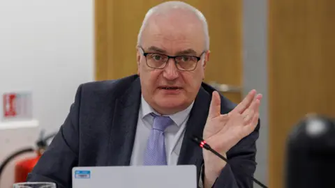 A man with white hair, glasses, navy blazer, white shirt and purple tie, he has his left hand raised.