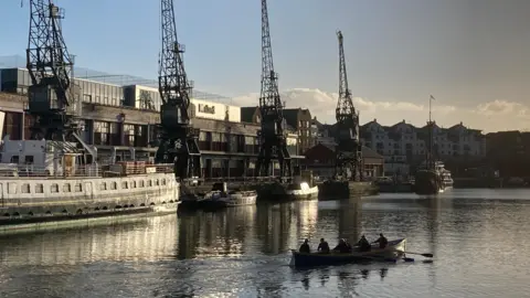 Triassic Trekker People are rowing a boat in water in a harbour. Beside it are tall cranes and a large building which says M Shed, and a ship.