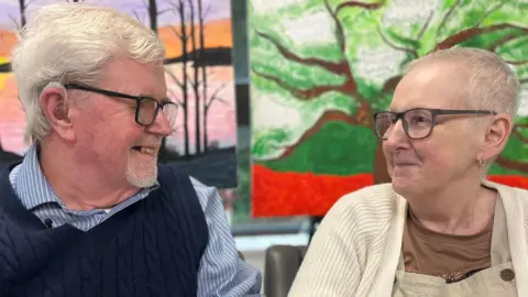 Sally and Kevin are looking at each other and smiling. Kevin is wearing a blue and white striped shirt and a navy knitted tank top. Sally is wearing a cream cardigan, brown t-shirt and light brown dungarees. Both have short white hair and glasses.