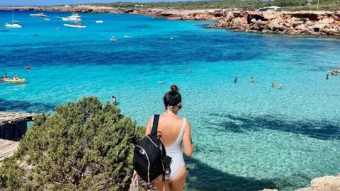 Monica Stott Monica sedang berjalan ke pantai dengan pakaian renang dengan ransel hitam dan baju renang biru muda. Punggungnya menghadap kamera dan di depannya kita bisa melihat perairan biru cerah. 