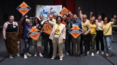 BBC  Layla Moran in a cream suit and white shoes, with about 12 supporters cheering 