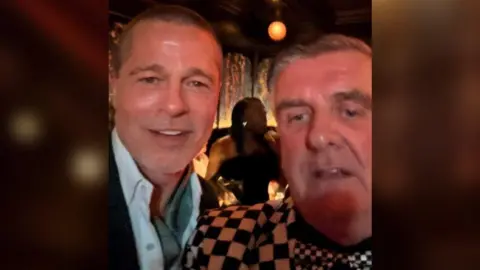A selfie of Brad Pitt, who is wearing a blue shirt which is open, who is standing on the left smiling at the camera. On the right is Martin Donnelly who is looking towards the camera smiling, and is wearing a black and white chequered suit jacket. 