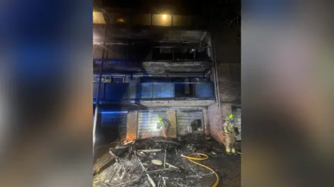 A block of flats severely damaged by fire. Firefighters an be seen assessing the damage. The fire is out. 