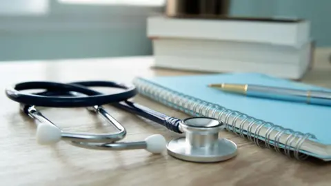 Stethoscope, pen, notebook and books on a wooden desk. Sunlight filters through the window and light curtain in the background.